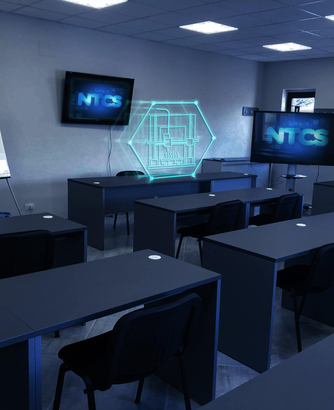 Viene mostrata un'aula moderna attrezzata con banchi e sedie. Due grandi schermi mostrano il logo "NTCS", uno sulla parete e l'altro su un supporto. Al centro, un'icona olografica con elementi elettronici aggiunge un'atmosfera high-tech alla stanza, ideale per corsi di stampa 3d a Napoli.